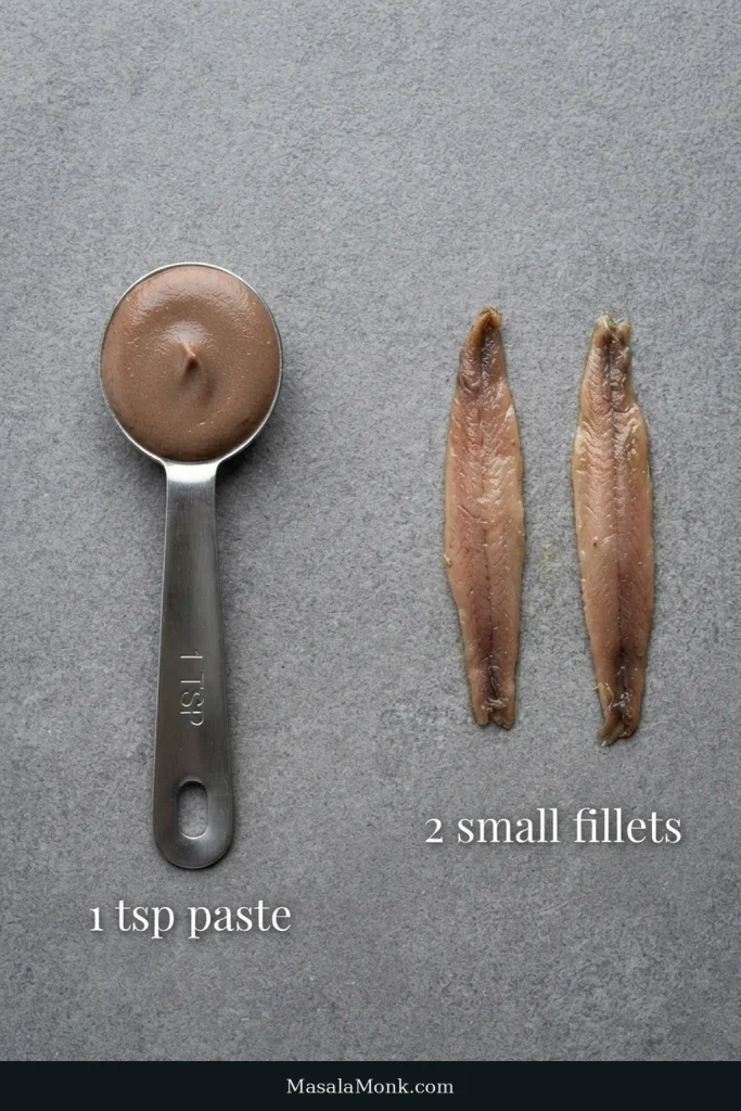 A measuring teaspoon of anchovy paste sits beside two small brown oil-packed anchovy fillets.
