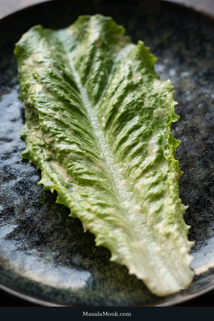 A crisp romaine leaf carries a thin, irregular coating of Caesar dressing, with the central rib and veins still visible.