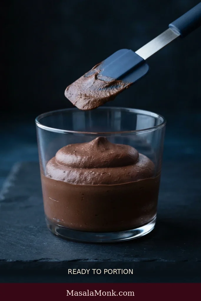 Freshly folded chocolate mousse rests in a clear serving glass while a spatula is held separately above it.