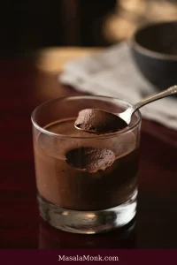 A clear glass of chocolate mousse with a small bite on a silver spoon, revealing the creamy surface left by the spoon.