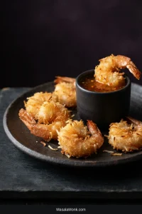 Plate of golden coconut shrimp with crisp shredded-coconut and panko coating beside a glossy dipping sauce and lime wedges.