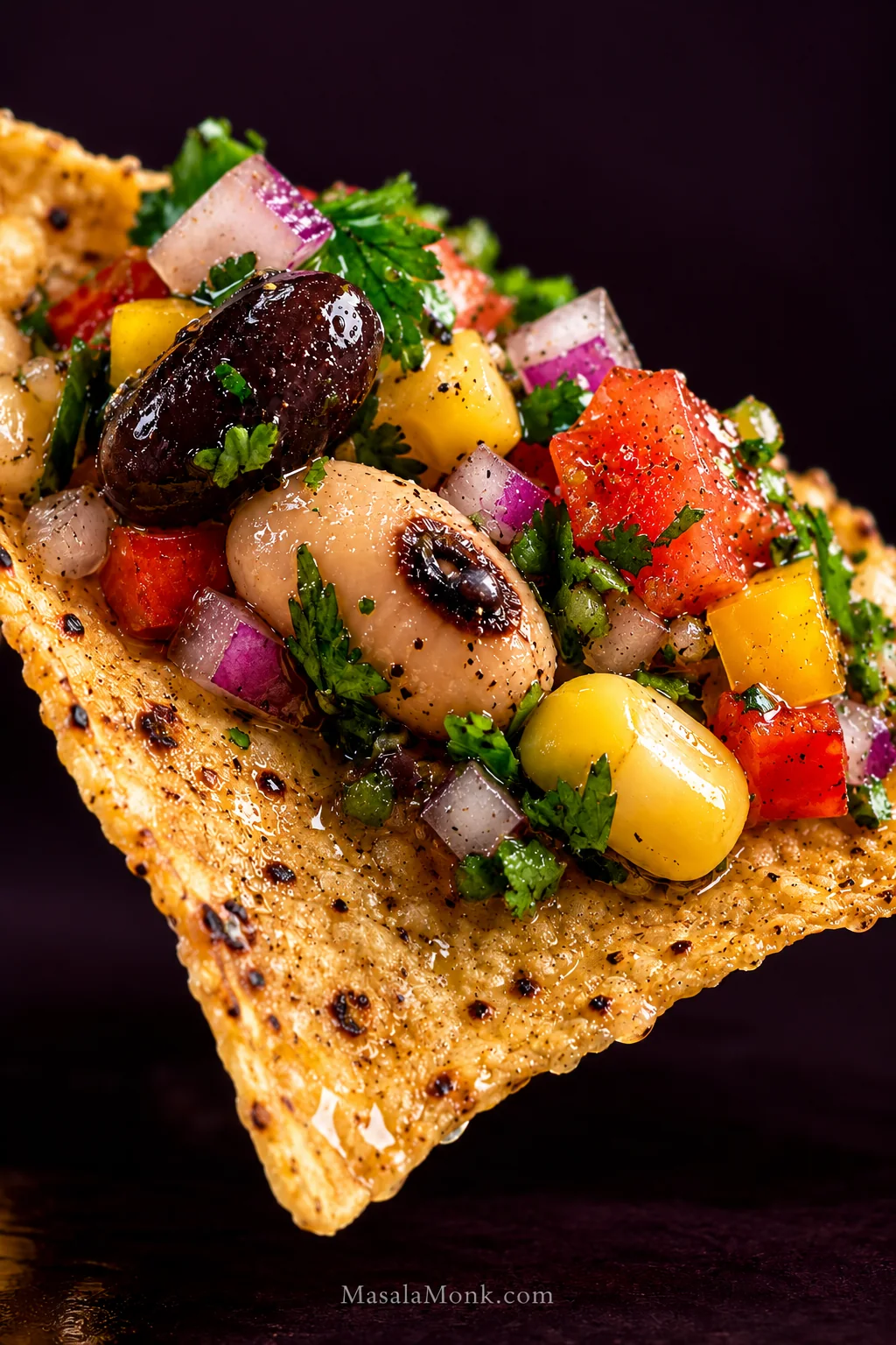 Extreme close-up of a tortilla chip topped with black-eyed peas, a black bean, corn, tomato, red and yellow bell pepper, red onion, cilantro and a light vinaigrette sheen.