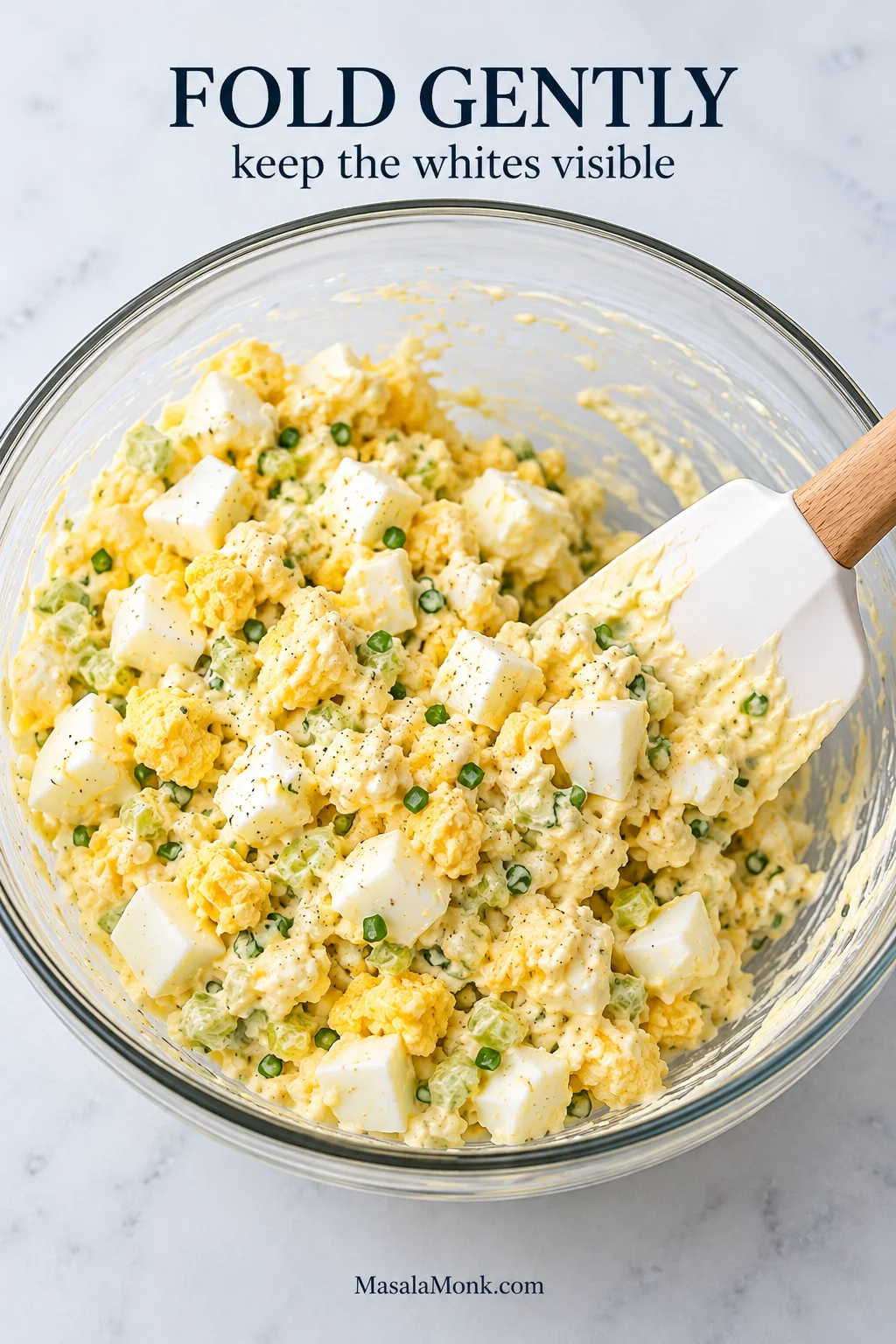 Glass bowl of partly mixed egg salad with large visible egg-white pieces and finely chopped green vegetables being gently folded with a spatula.
