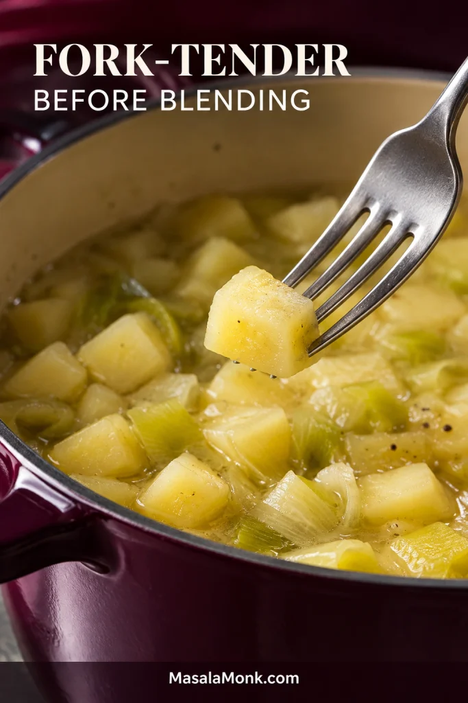 Fork piercing a tender half-inch piece of cooked Yukon Gold potato in a pot with softened leeks and broth before blending.