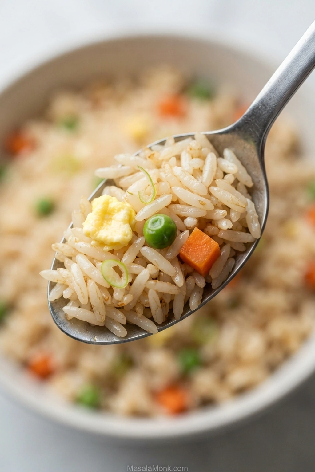 Close-up spoonful of pale golden fried rice with separate grains, a small egg piece, diced carrot, one green pea and thin scallion slices.