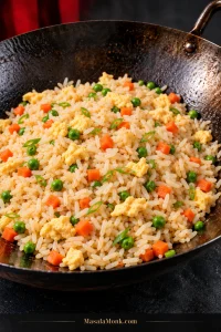 Finished fried rice in a seasoned carbon-steel wok with pale golden rice, small pieces of scrambled egg, diced carrots, green peas and sliced scallions.