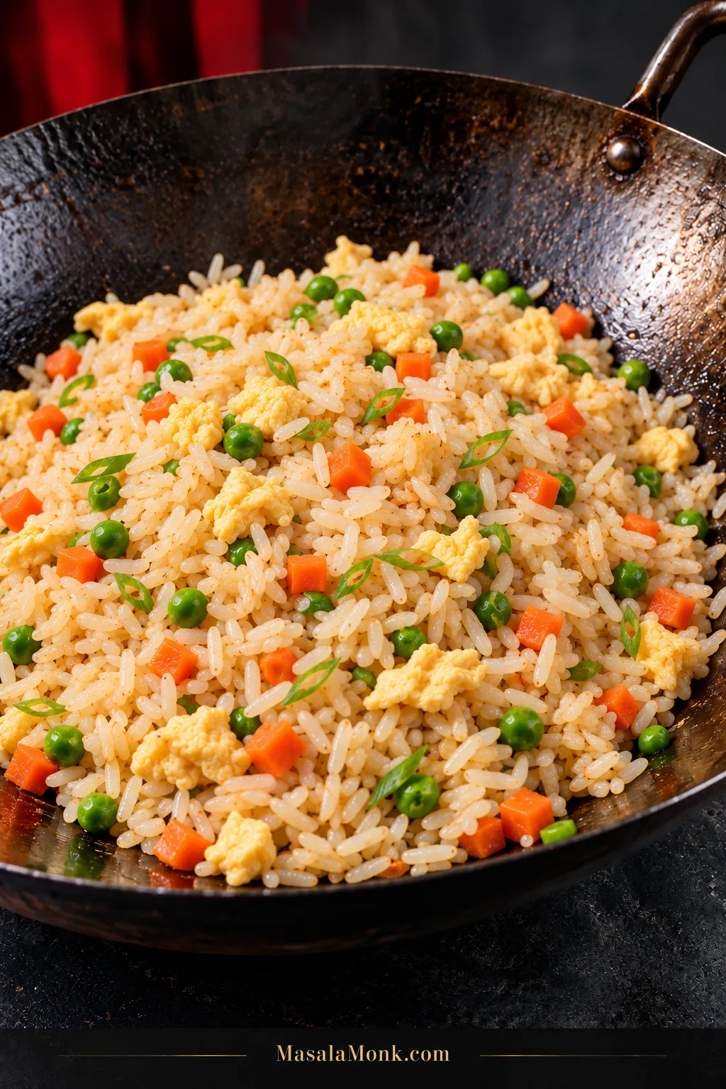 Finished fried rice in a seasoned carbon-steel wok with pale golden rice, small pieces of scrambled egg, diced carrots, green peas and sliced scallions.