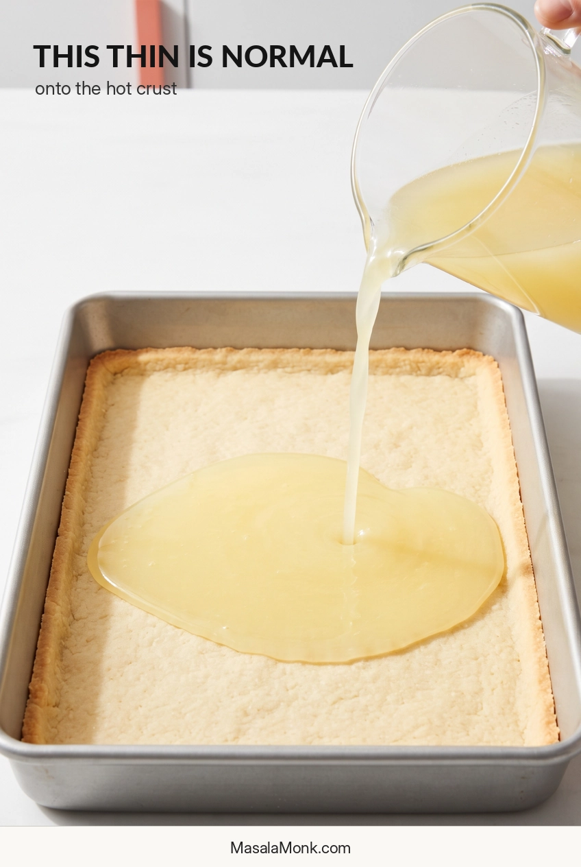 Thin pale-yellow lemon filling pouring from a clear pitcher onto a flat par-baked shortbread base in a rectangular metal baking pan.