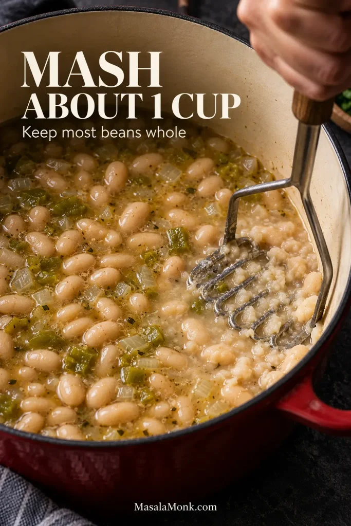 Potato masher crushing some cooked white beans against the side of a pot while most of the beans remain whole in the broth.