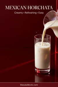 Warm-white Mexican horchata pouring from a clear pitcher into a tall glass filled with clear ice against a deep ruby background.