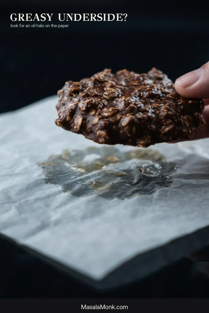 A chocolate oatmeal no-bake cookie lifted from paper with a translucent oil halo visible underneath.