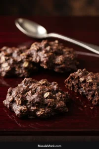 Four dark chocolate peanut butter oatmeal no-bake cookies on a deep red surface, including one broken cookie showing a dense oat-studded center.