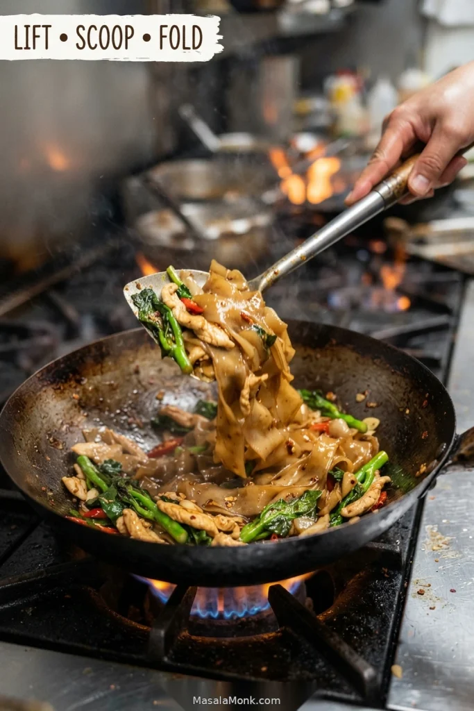Pad Kee Mao noodles being lifted and folded in a hot wok with chicken, green vegetables and red chillies.