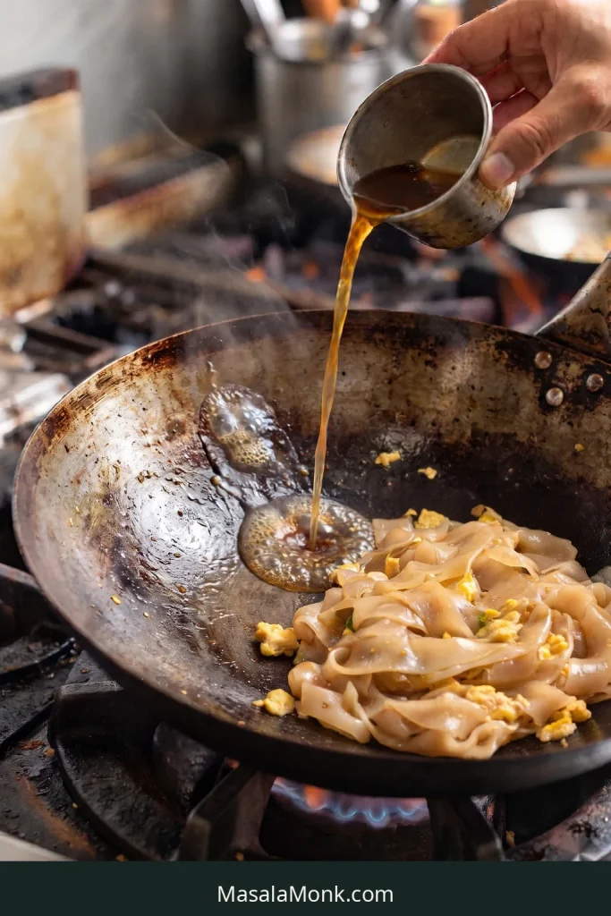 Thin amber-brown Pad See Ew sauce poured onto exposed hot wok metal beside pale wide rice noodles and egg.