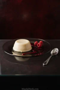 Unmolded vanilla panna cotta on a dark plate with raspberry coulis, fresh raspberries and a silver spoon.
