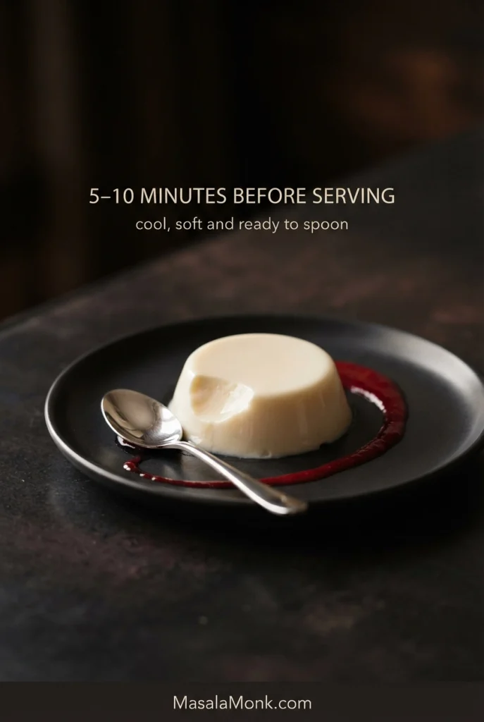 Partly spooned vanilla panna cotta on a dark plate with raspberry coulis and a silver spoon.