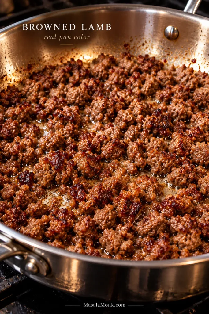 Small irregular ground lamb crumbles browning in a stainless-steel skillet, with darker caramelized patches on meat touching the pan.