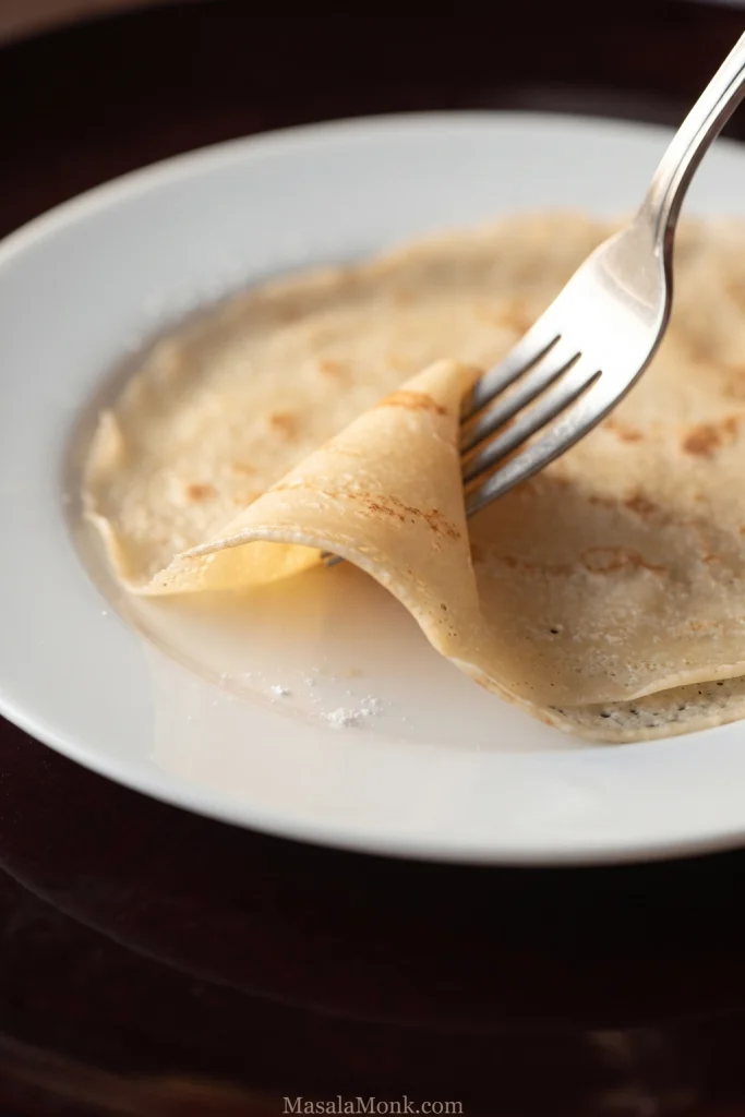 Silver fork gently lifting a thin folded crepe from a white plate, showing the pale-golden crepe bending in soft flexible layers.
