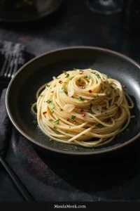 Shallow white bowl of spaghetti aglio e olio with glossy strands, thin pale garlic slices, parsley, and small red chile pieces on a dark graphite surface.