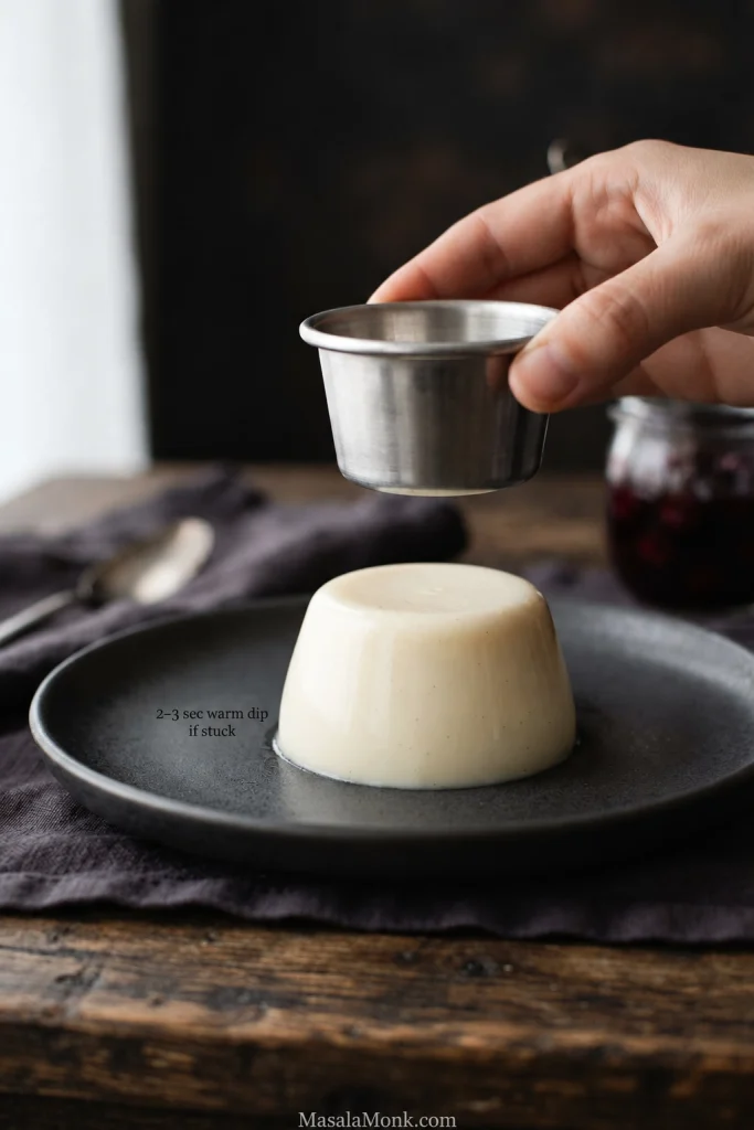 Hand lifting a metal mold from an unmolded panna cotta, with a note about a 2 to 3 second warm-water dip if stuck.