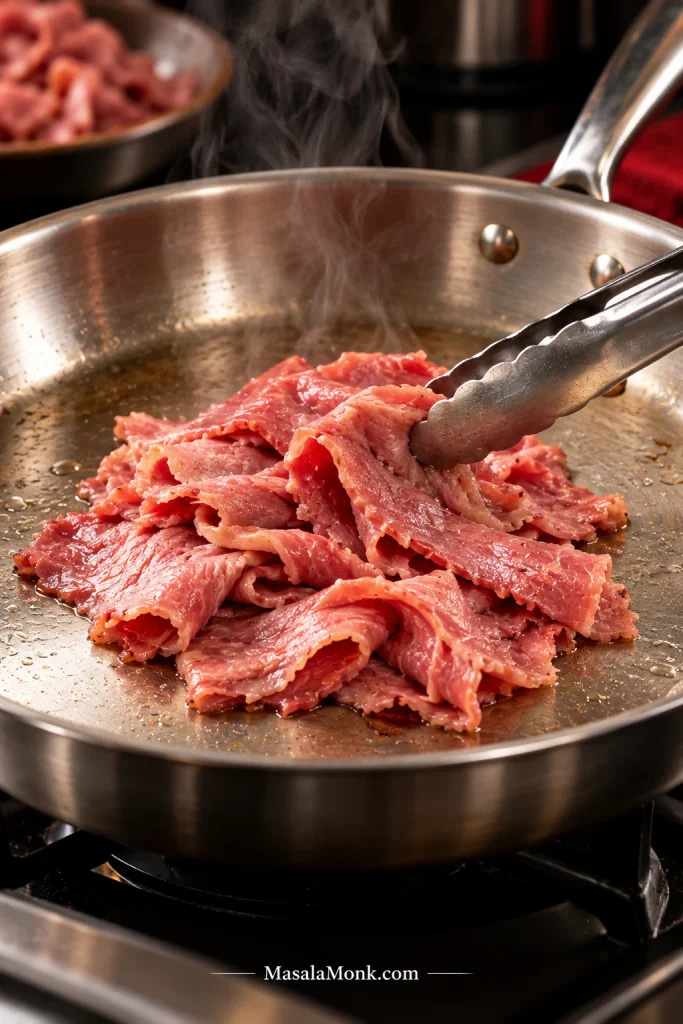 Thin slices of pink corned beef warming in loose folds in a stainless-steel skillet while metal tongs lift and separate the meat.
